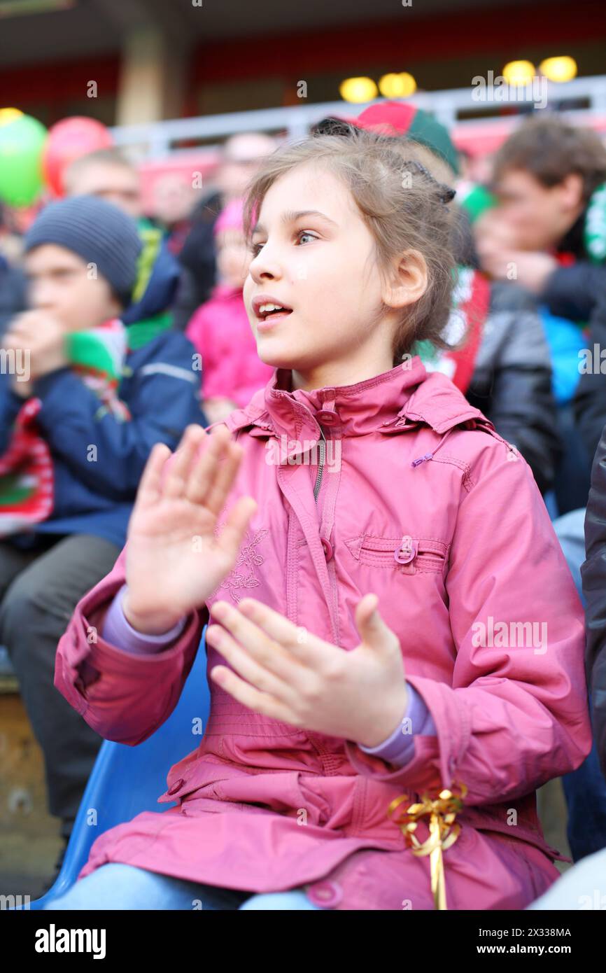 Little girl in a pink jacket applauds among fans at a sports stadium ...