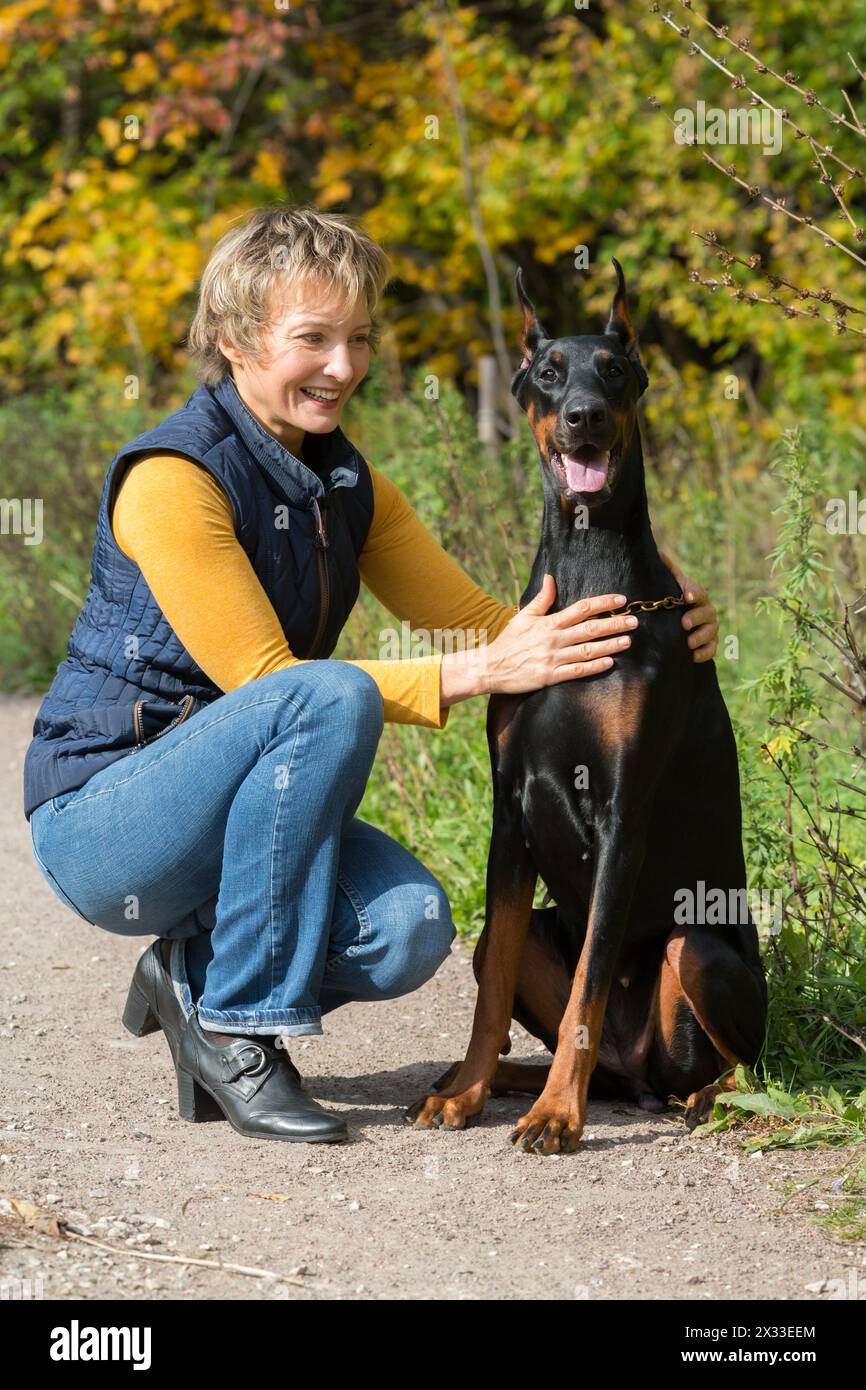 Smiling woman at beige polo-neck is hugging dobermann in a park. Stock Photo