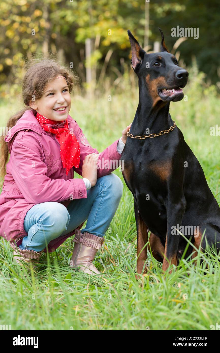 Little girl is smiling and sitting on the grass near a dobermann. Stock Photo