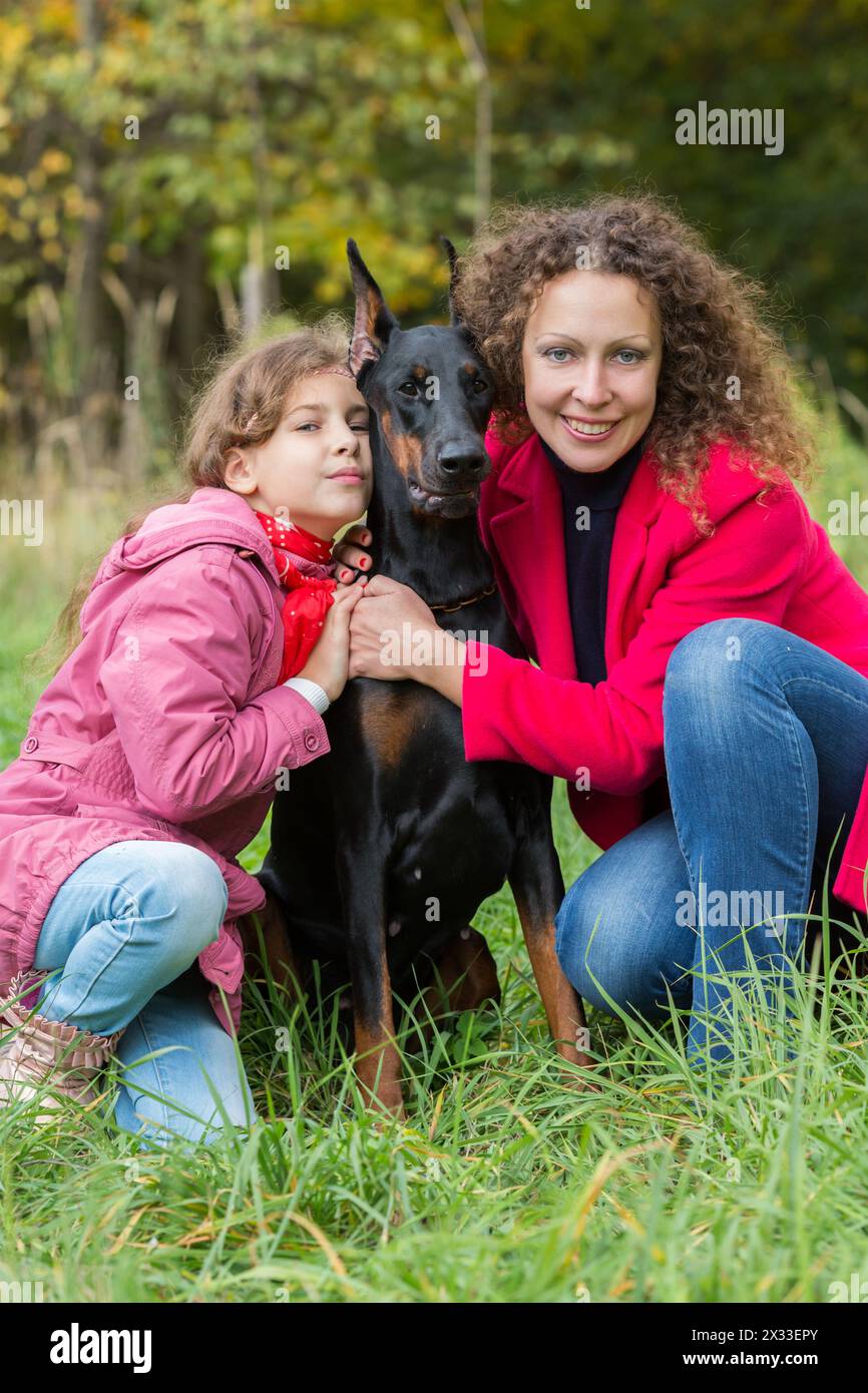 Little curly girl and her mother are sitting near dobermann in park. Stock Photo