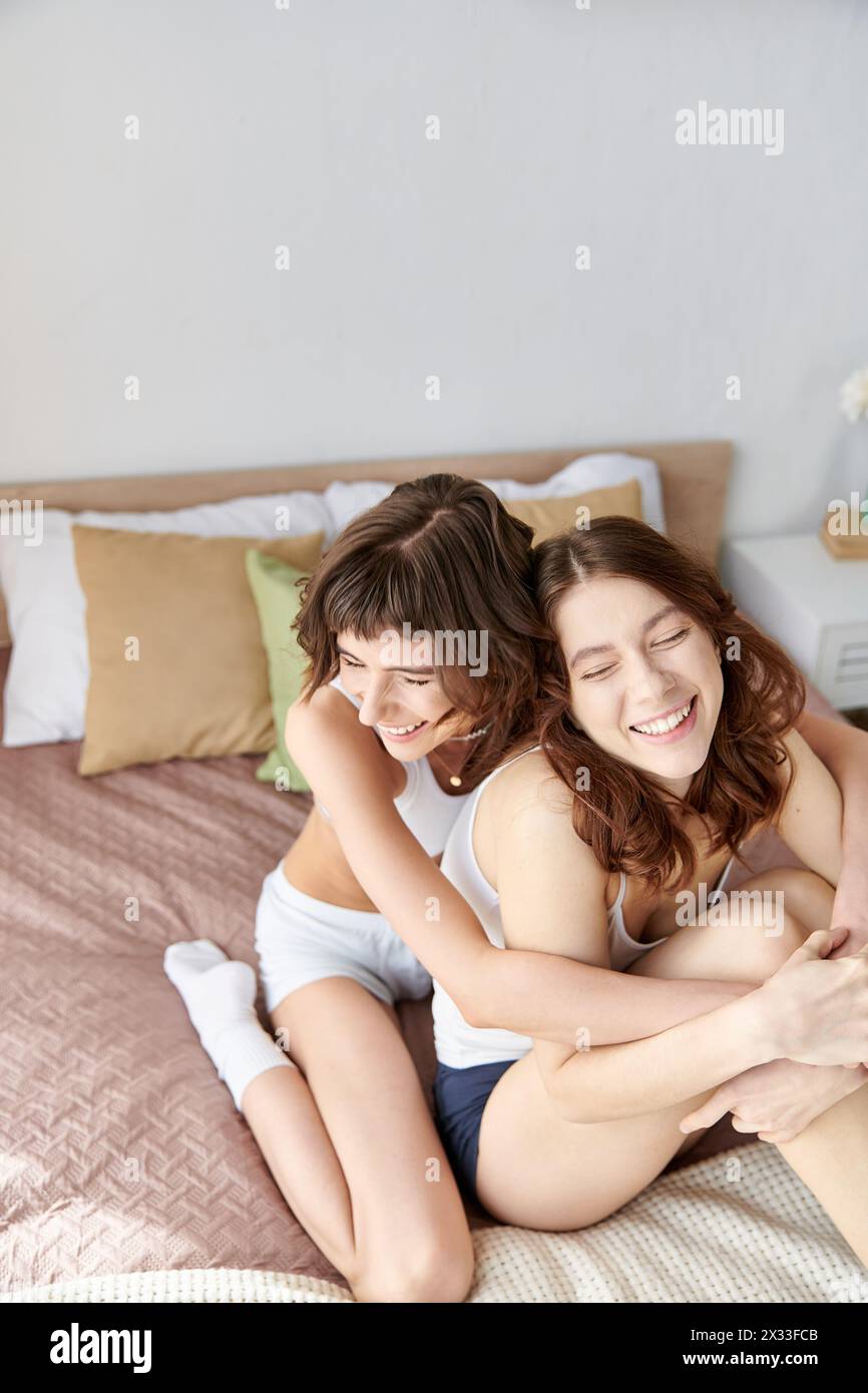 Two women in cozy attire sit on a bed, hugging each other warmly. Stock Photo