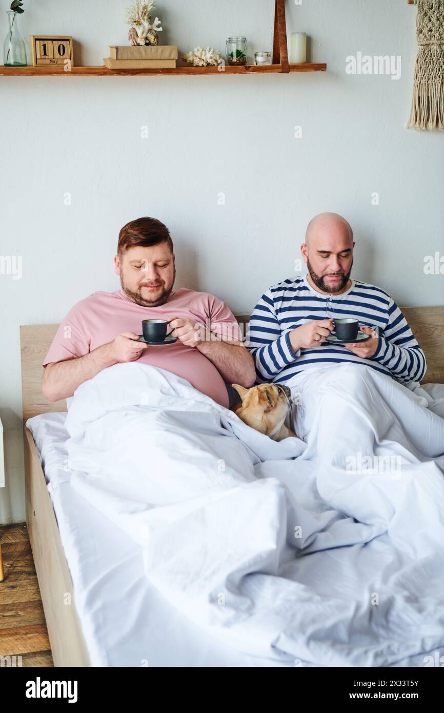 A gay couple sits together on a bed, accompanied by their French bulldog. Stock Photo