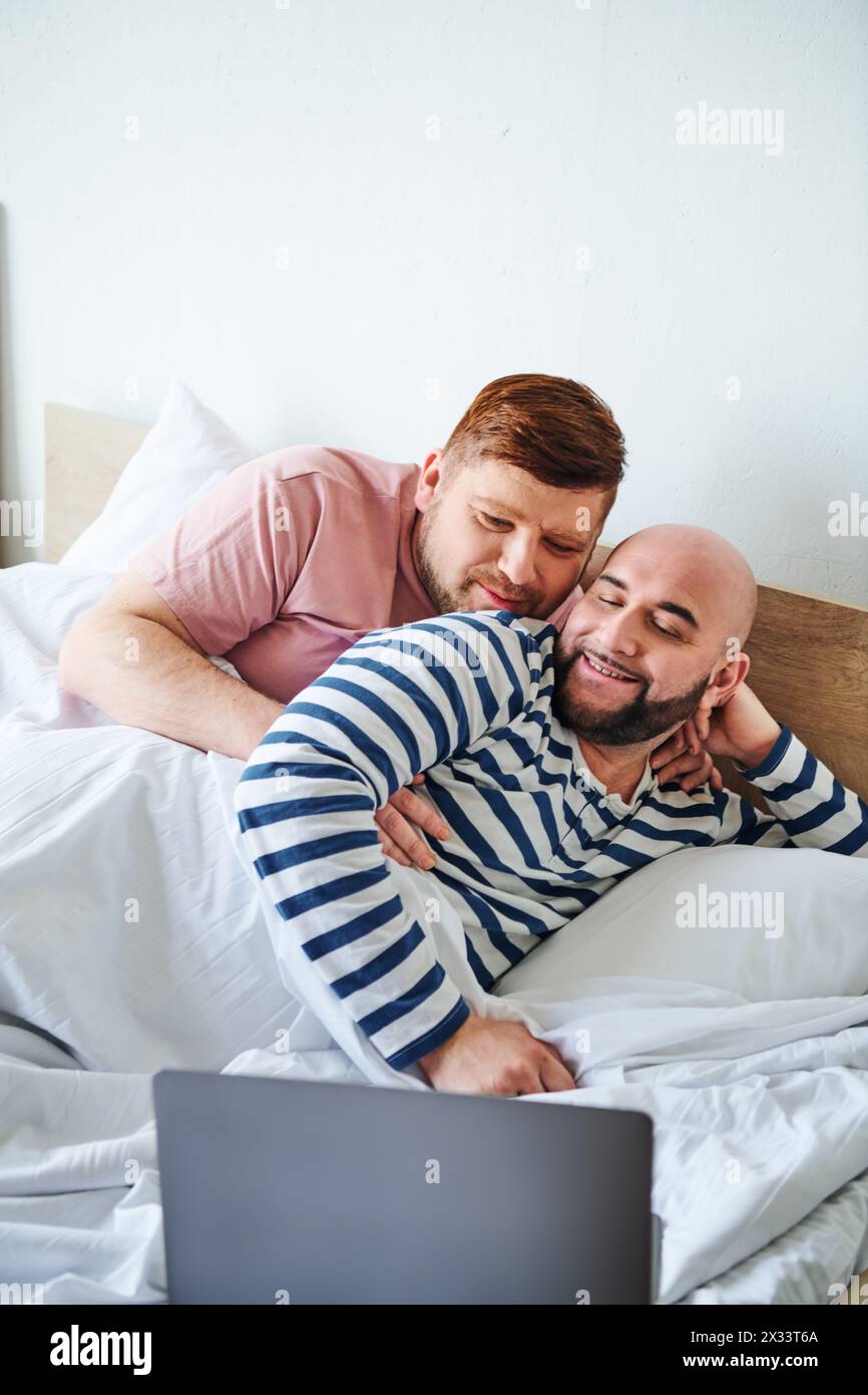 Two men cozy up in bed while using a laptop. Stock Photo