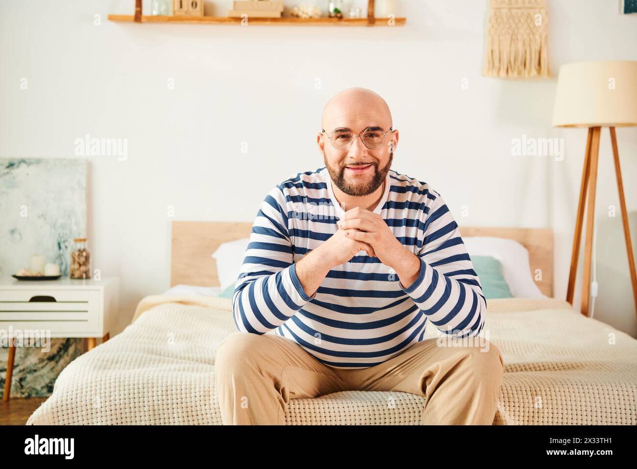 A man in glasses sitting on a bed in a bedroom, deep in thought. Stock Photo