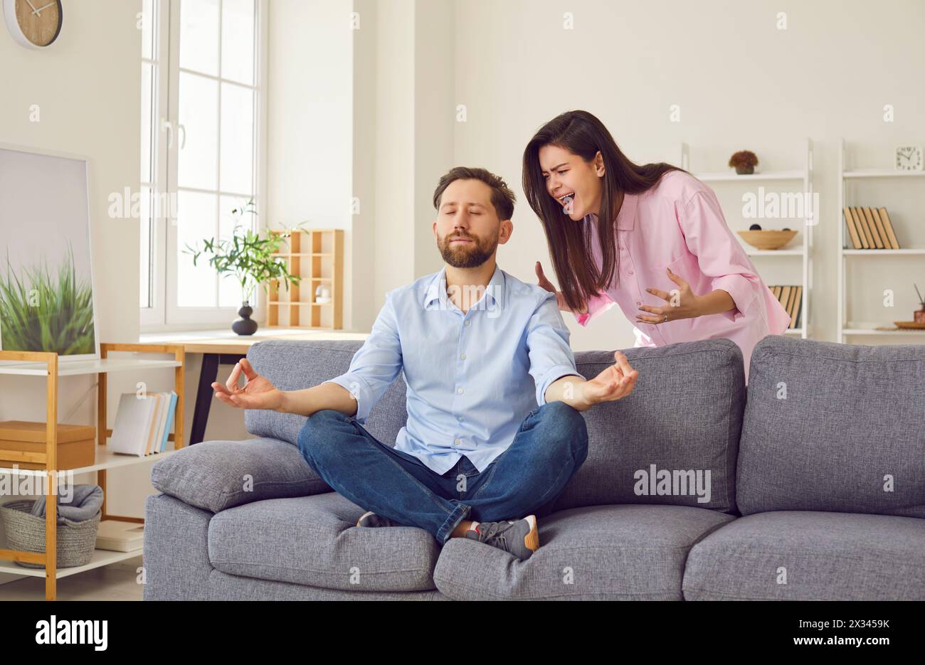Angry woman quarrels and shouts at man who is sitting in meditative position on sofa at home. Stock Photo