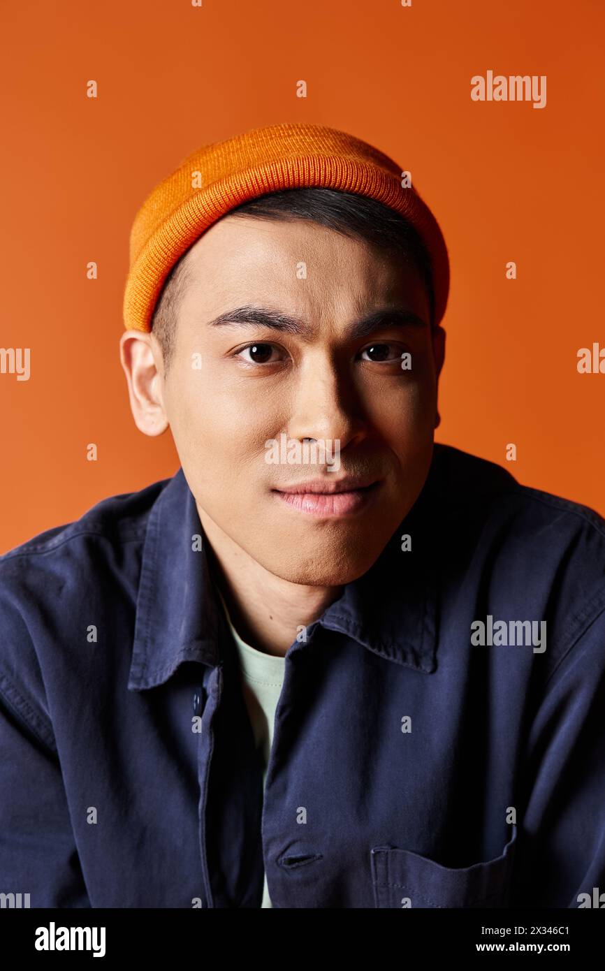 A handsome Asian man in a blue shirt and an orange hat stands confidently against an orange background in a studio. Stock Photo