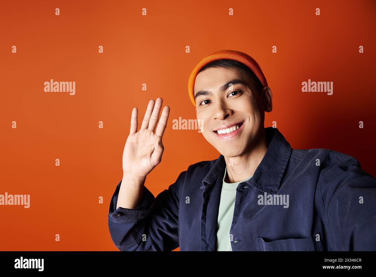 Handsome Asian man in blue jacket and orange hat confidently waving