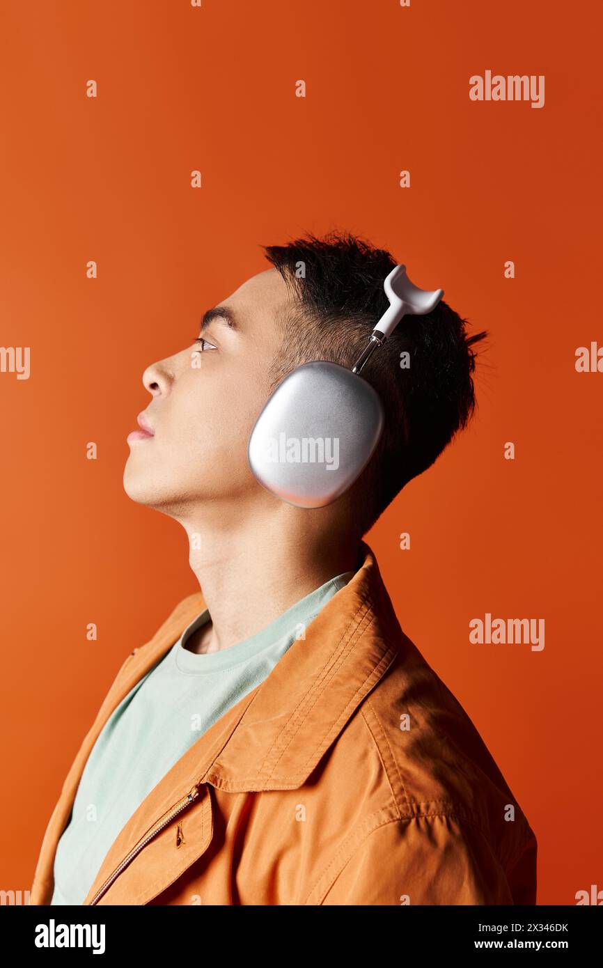 A handsome Asian man in stylish attire listening to music with headphones on his ears against an orange studio background. Stock Photo