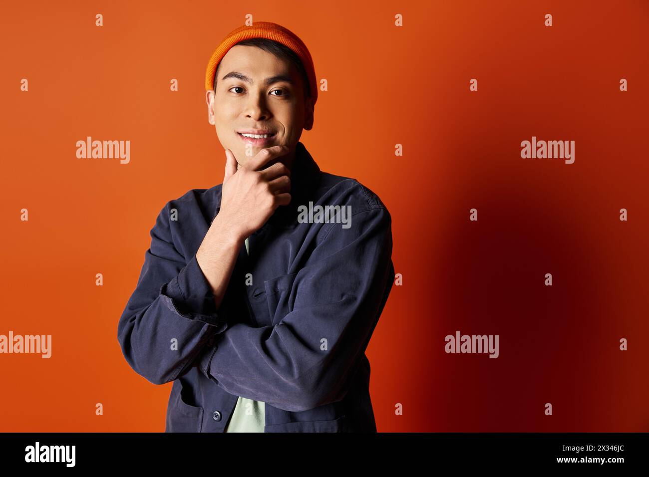 A handsome Asian man dressed in stylish attire stands confidently in front of an orange wall. Stock Photo
