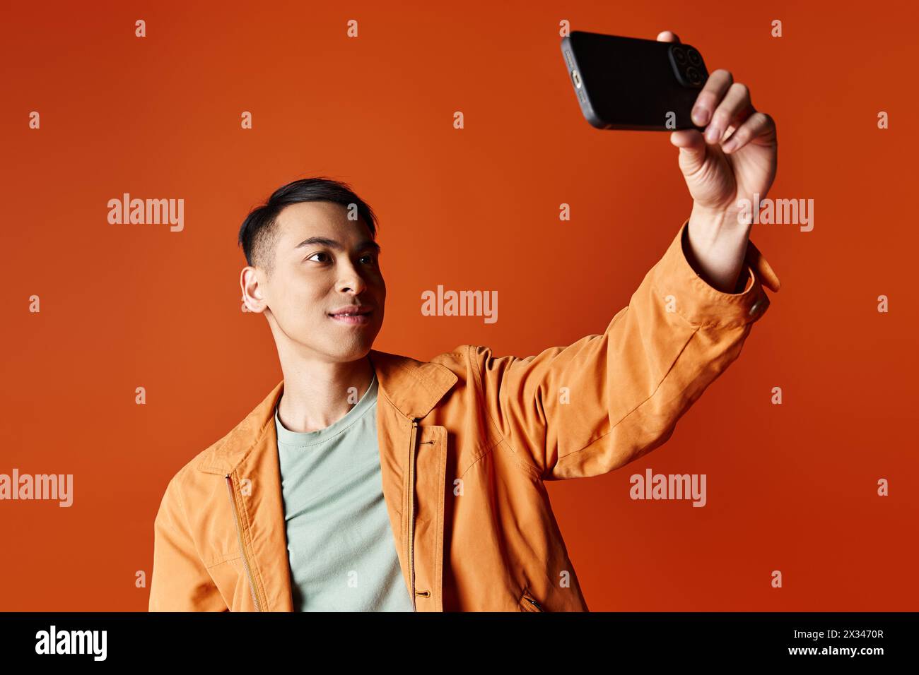 Handsome Asian man in stylish attire taking a picture with his cell phone against an orange studio background. Stock Photo