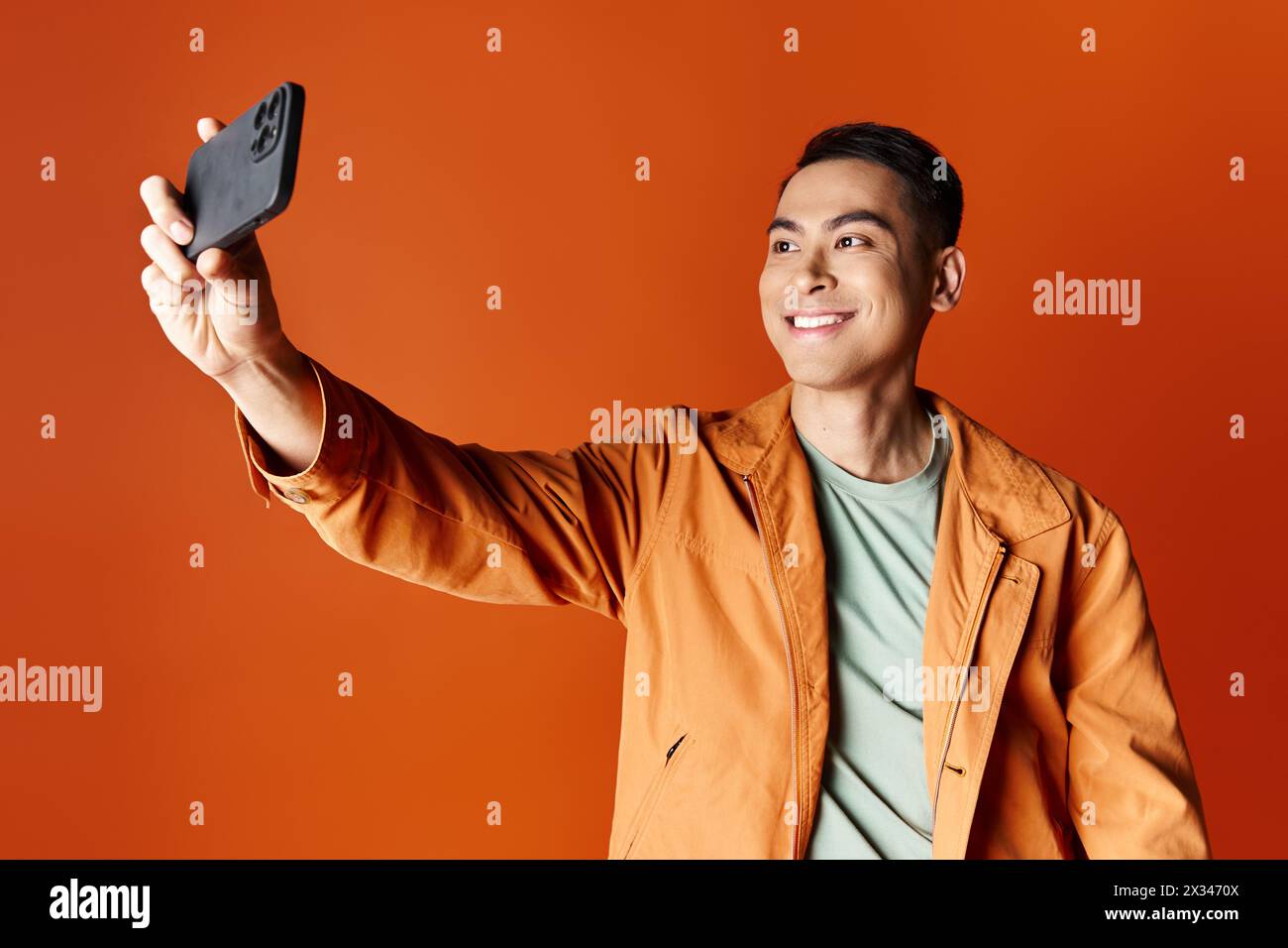 happy Asian man in stylish attire taking a selfie with his cell phone against an orange studio background. Stock Photo