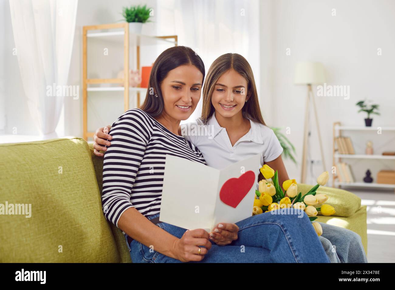 Young mother and daughter sitting on the sofa at home and reading a greeting card together. Stock Photo