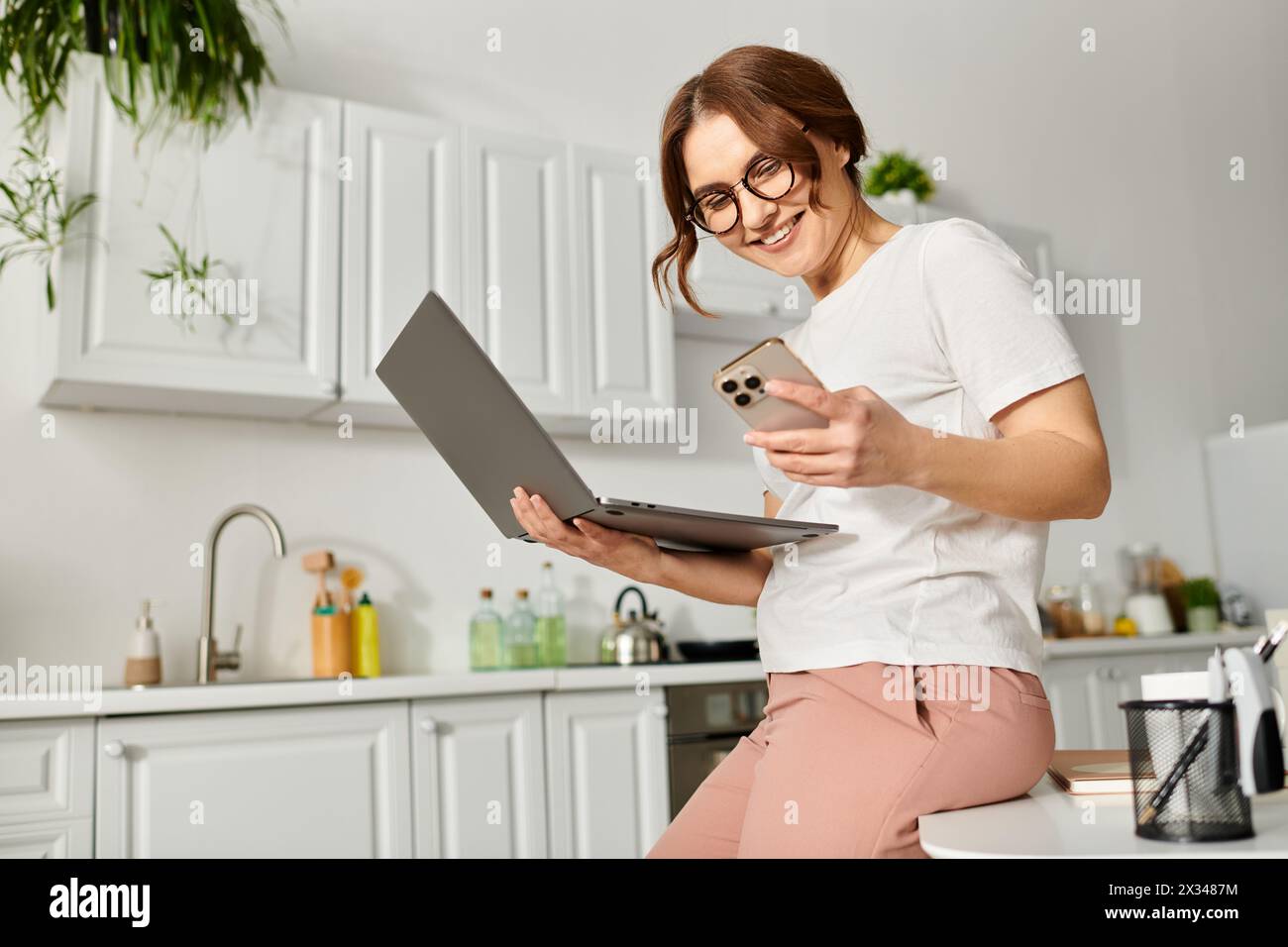 Middle-aged woman sits on counter, typing on laptop. Stock Photo
