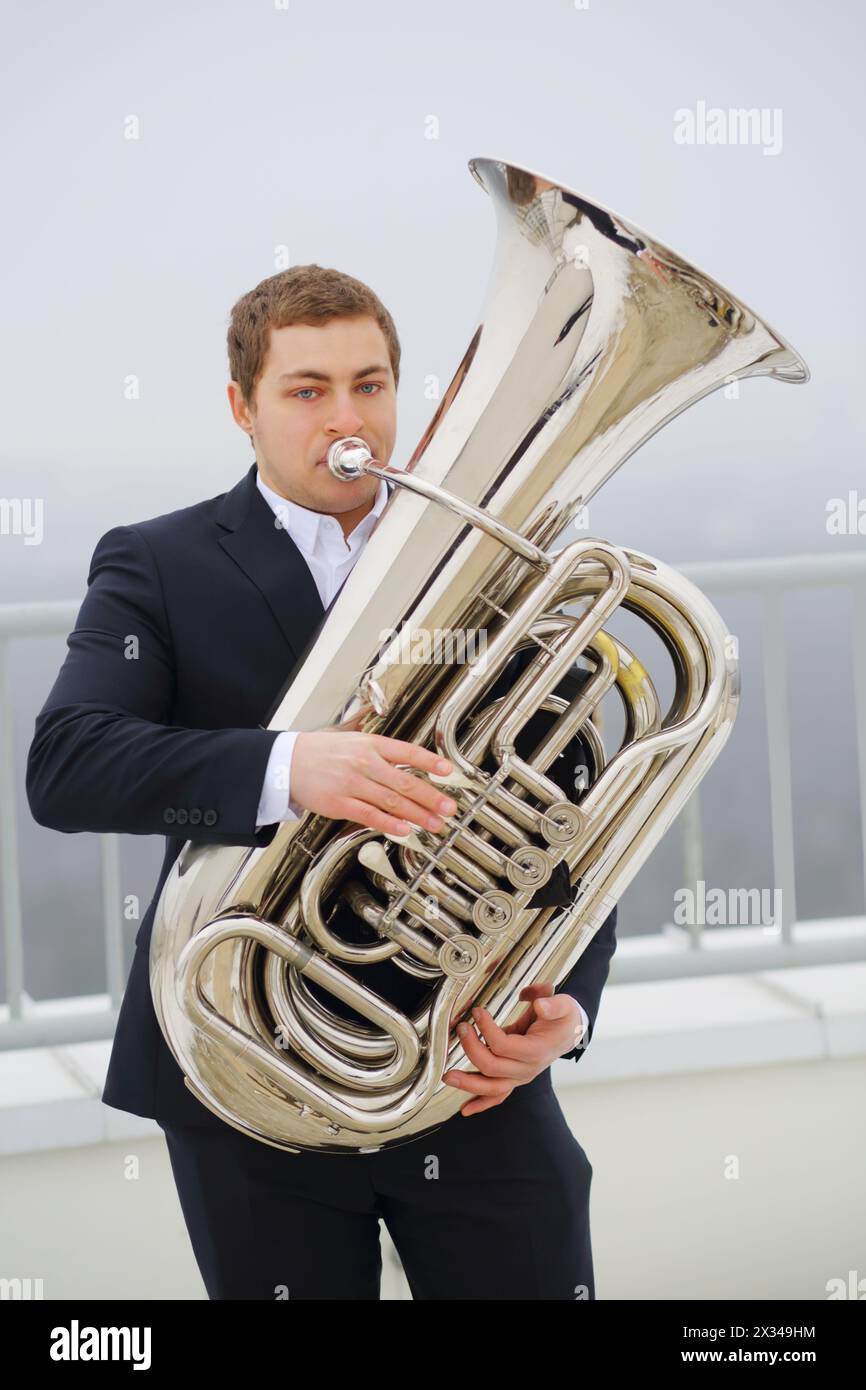 Man in black suit plays tuba on roof of tall building at winter day Stock Photo