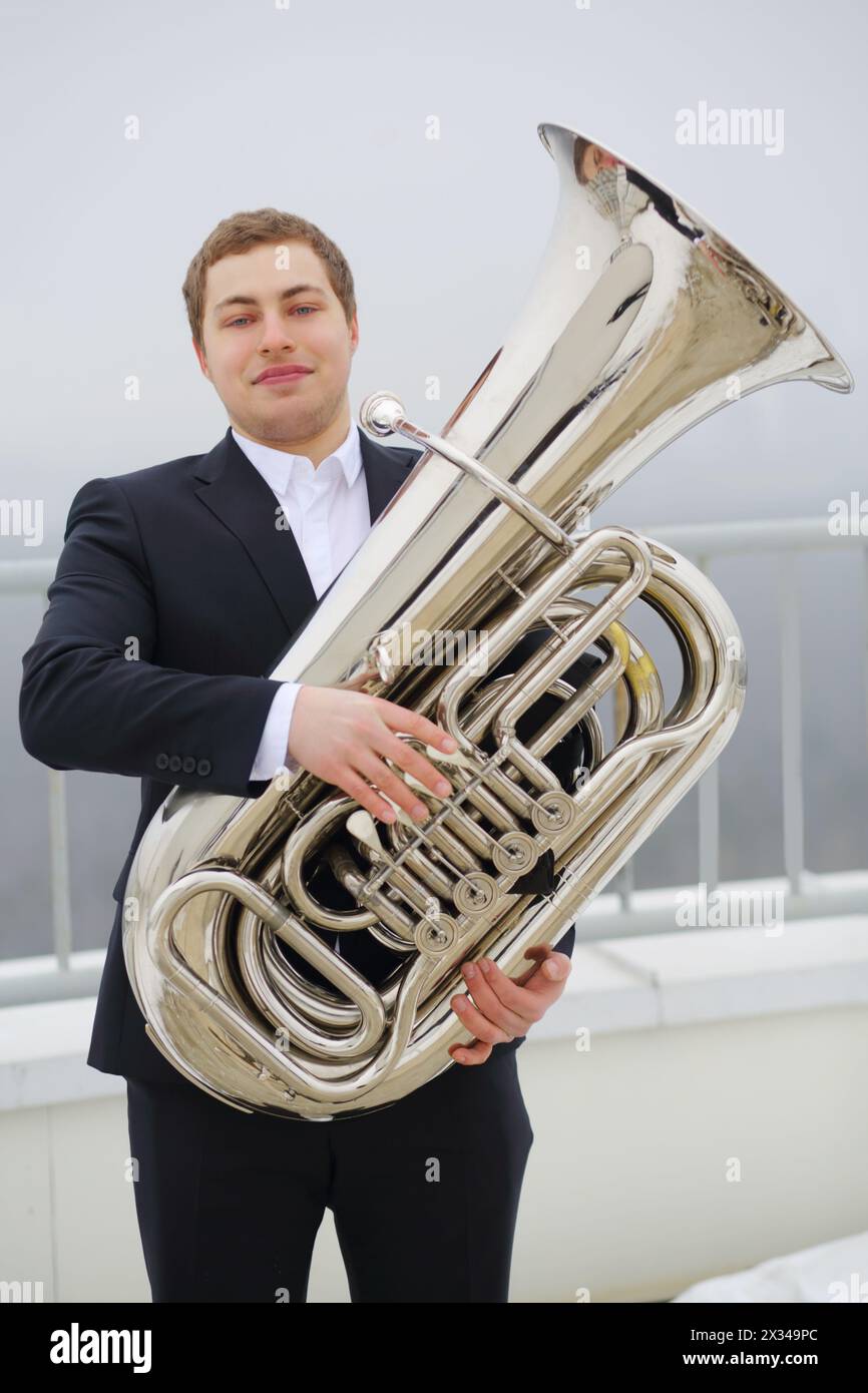 Man in black suit poses with tuba on roof of tall building at winter day Stock Photo