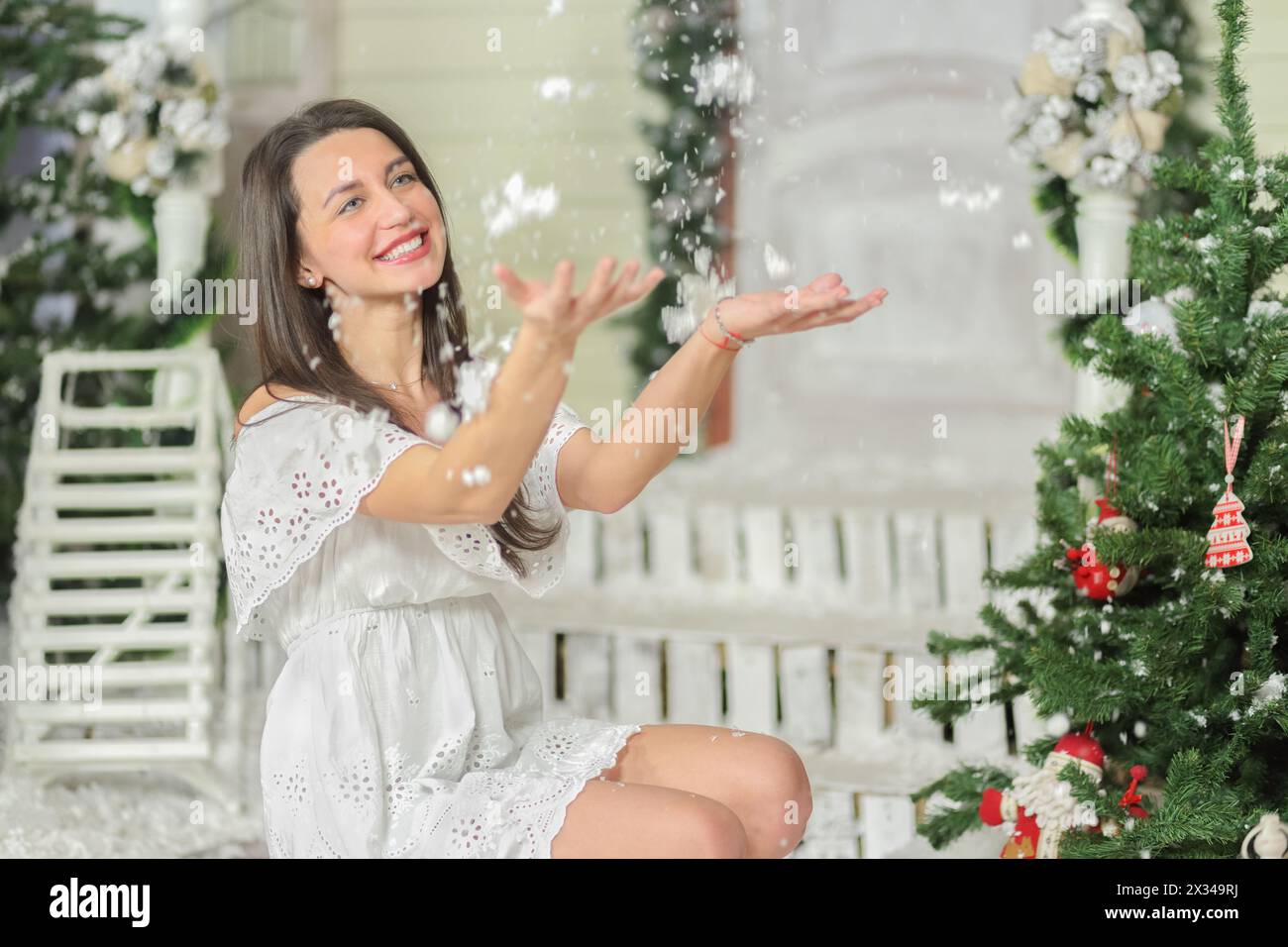 portrait of young woman sitting on squatting in courtyard of fake home and throws snow up, smiling. Christmas interior studio Stock Photo