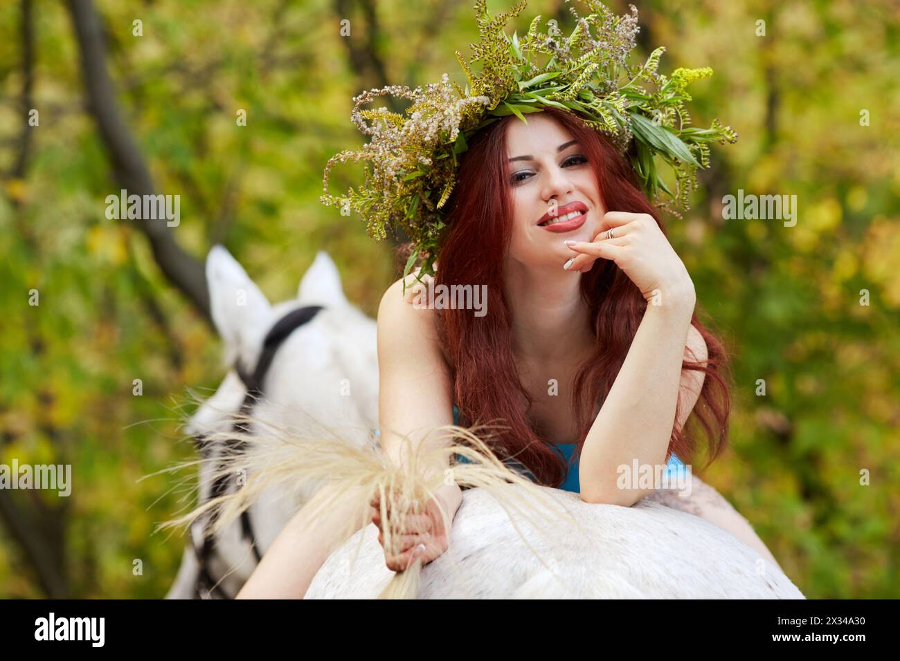 Young smiling red-haired woman with floral wreath on head lies on horseback in park. Stock Photo