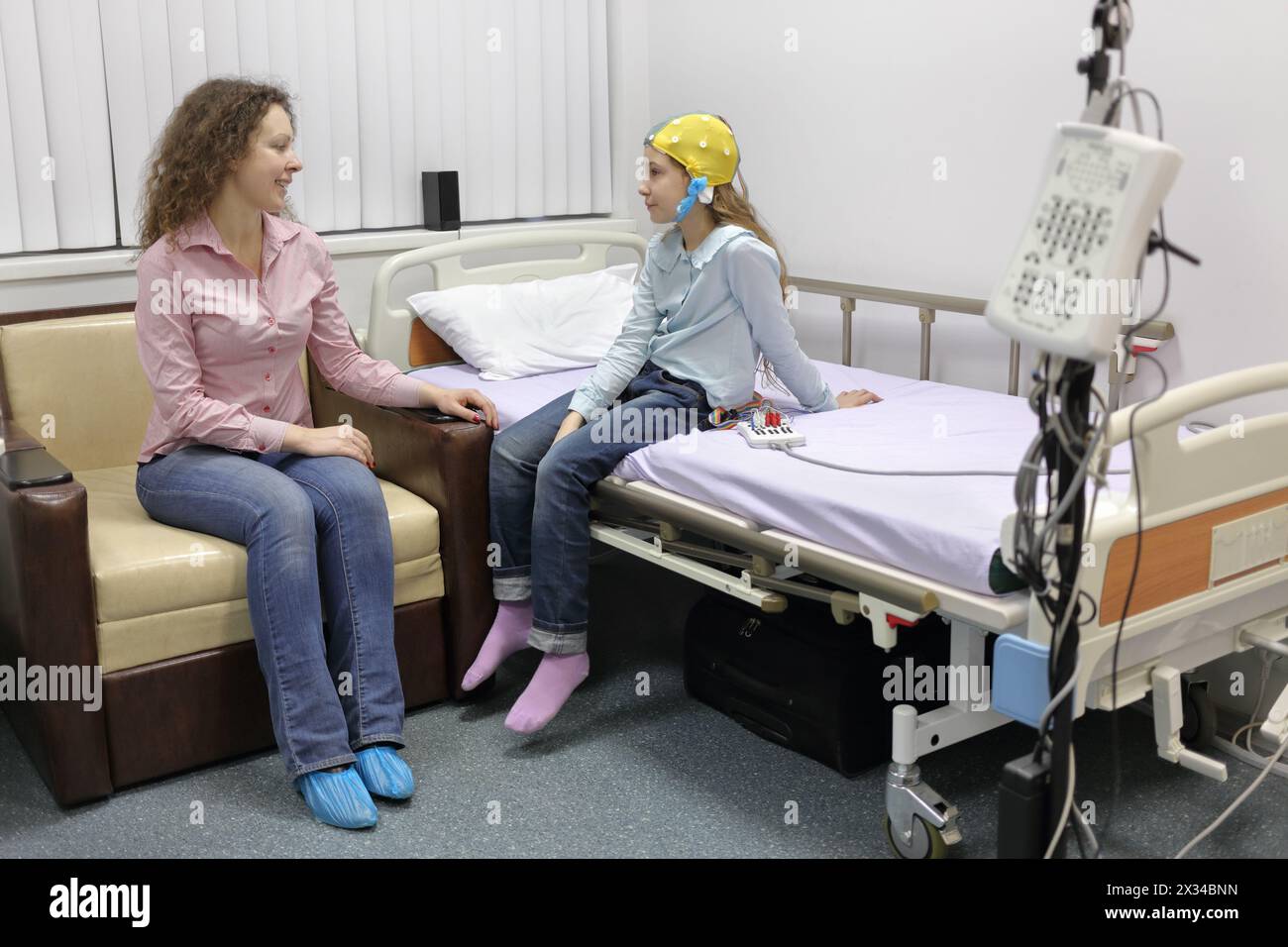 mother sitting beside her daughter at withdrawal procedure of electroencephalogram, talking to her Stock Photo