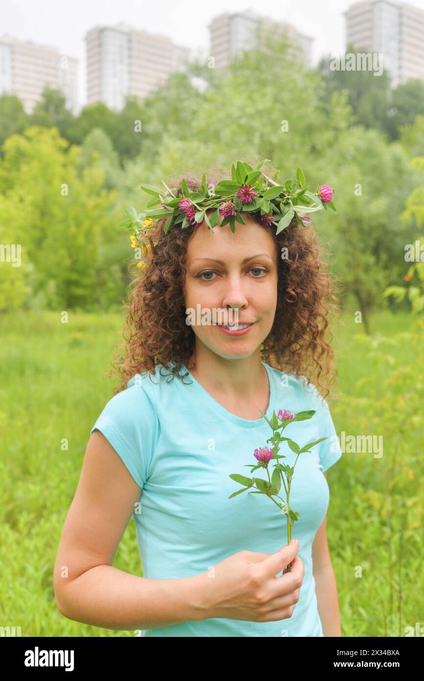 half length portrait of young woman with wreath of flowers urban wooded area near apartment complex, holding clover, smiling Stock Photo