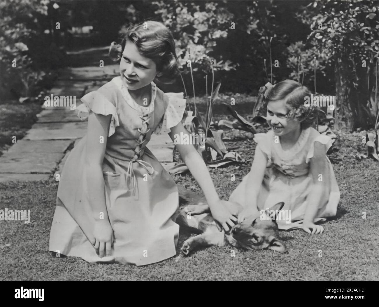 A young Princess Elizabeth, age 10, and Princess Margaret, age 6, are photographed together in the Royal Lodge, Windsor, dated 1936. This moment, captures the royal sisters during their early years, stroking one of their pet dogs. Stock Photo