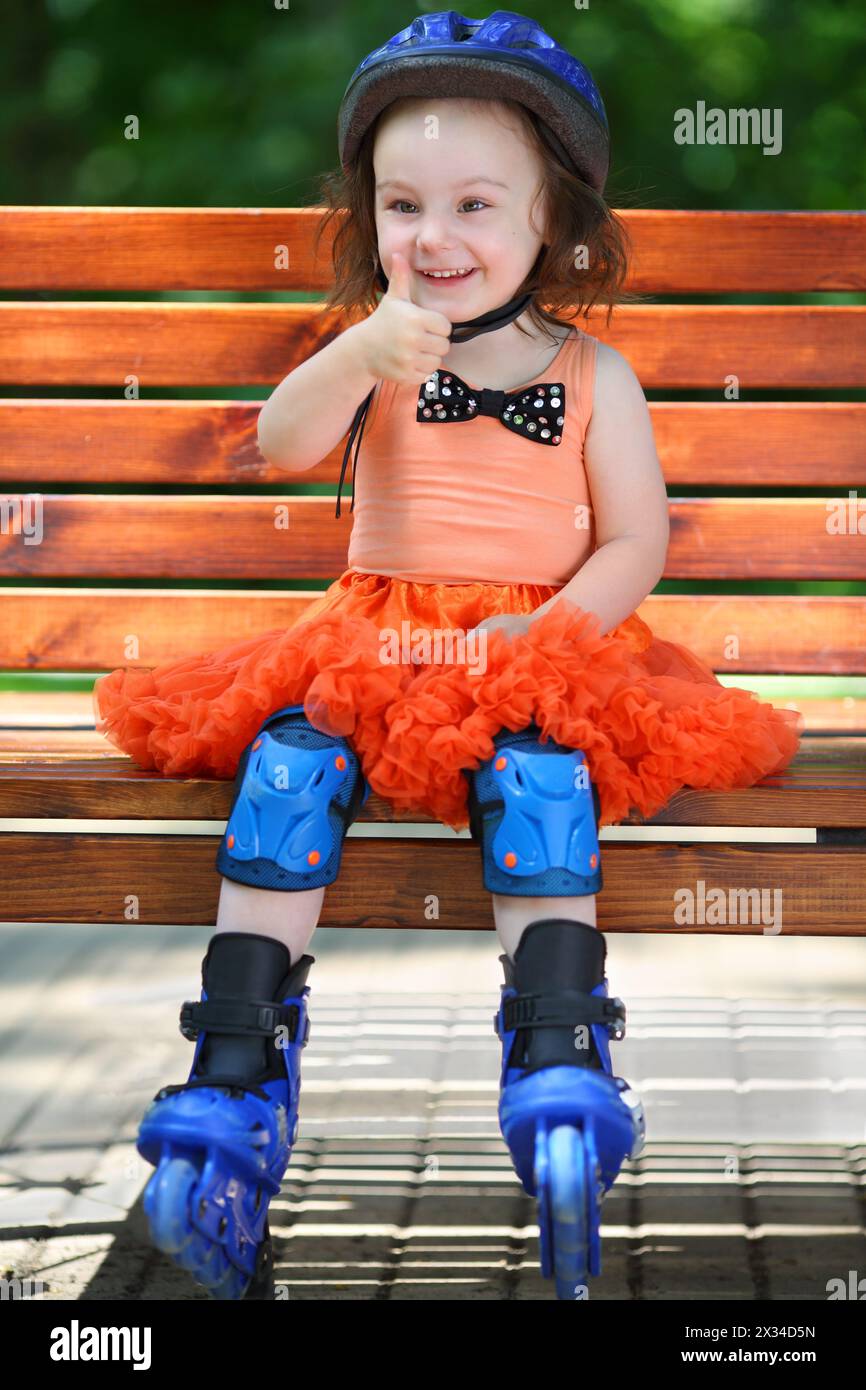 Girl in roller skates sits on bench and thumps up at summer day Stock Photo