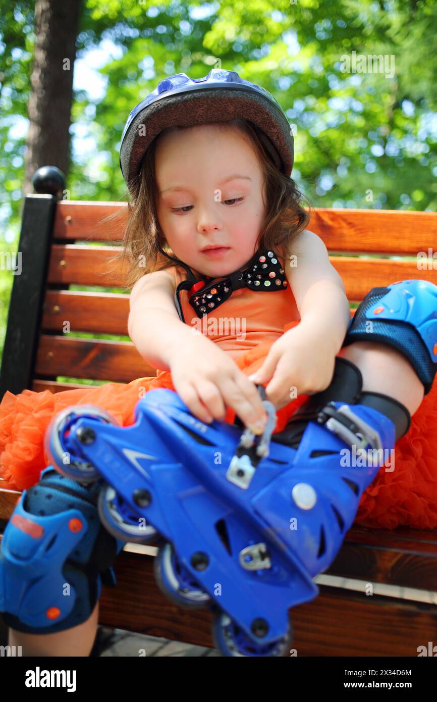 Little cute girl sitting on bench and removing roller skates at summer day Stock Photo