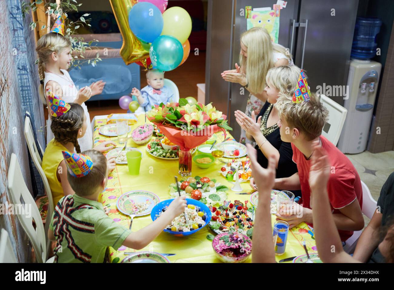 Seven adults and children sit at birthday table and congratulate little boy with applauses. Stock Photo