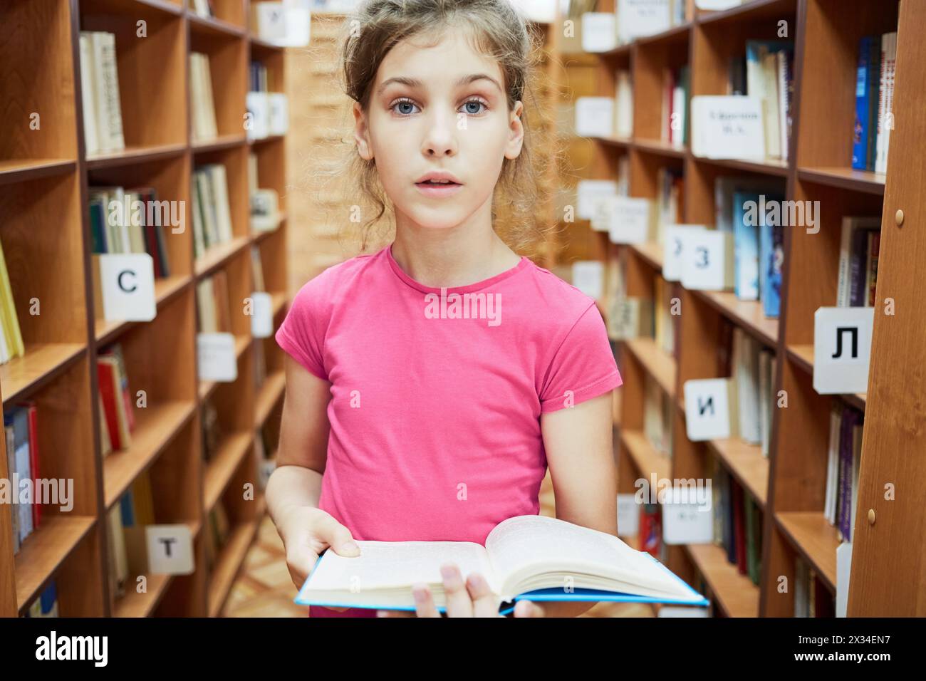 Teenage girl with wide open eyes stands holding book between bookcases