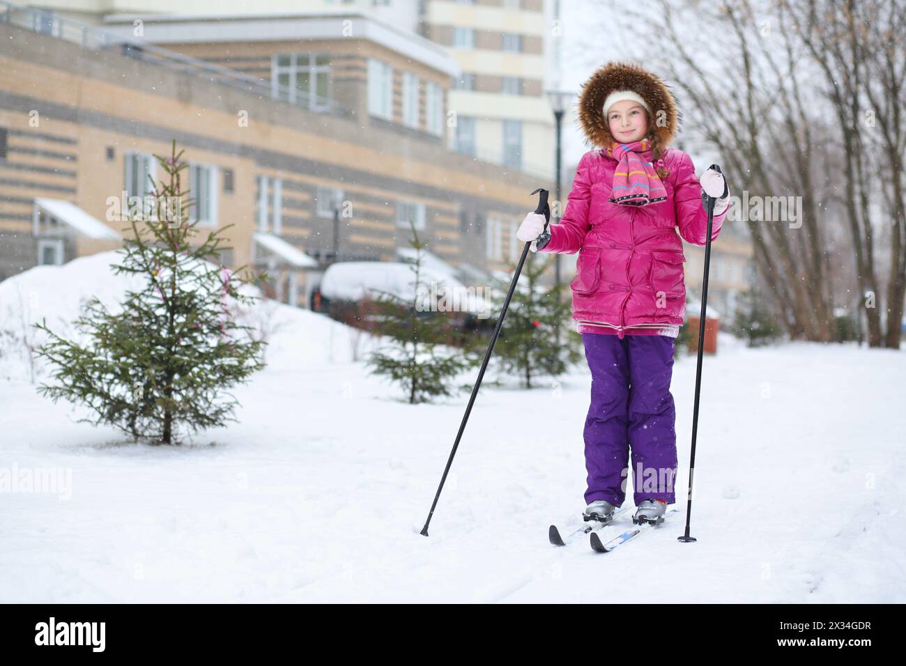 Little girl in a pink jacket with a hood skiing near the apartment complex Stock Photo