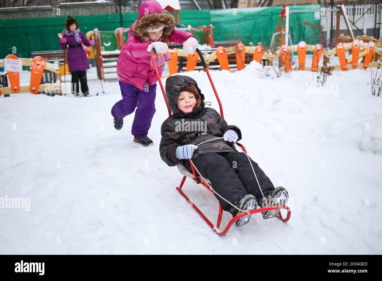 Happy girl rolling a little boy on a sled in front of the girl on skis in the winter playground Stock Photo