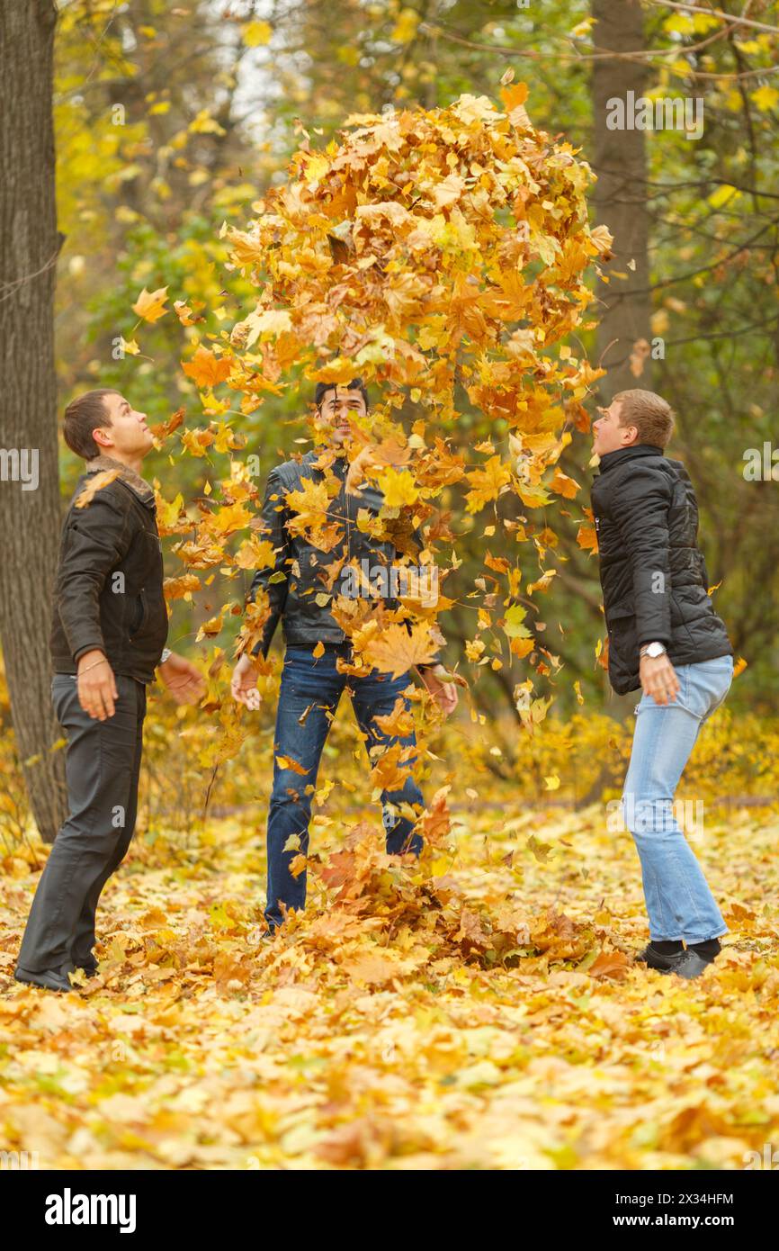Three men in black jackets pop-up yellow maple leaves in park Stock Photo