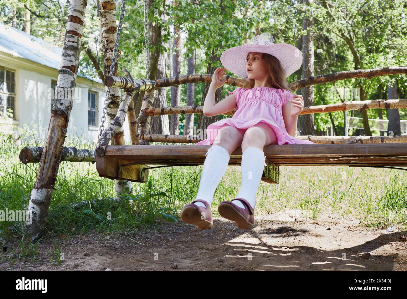 Little girl in pink dress and sunhat sits on wooden swings under trees ...