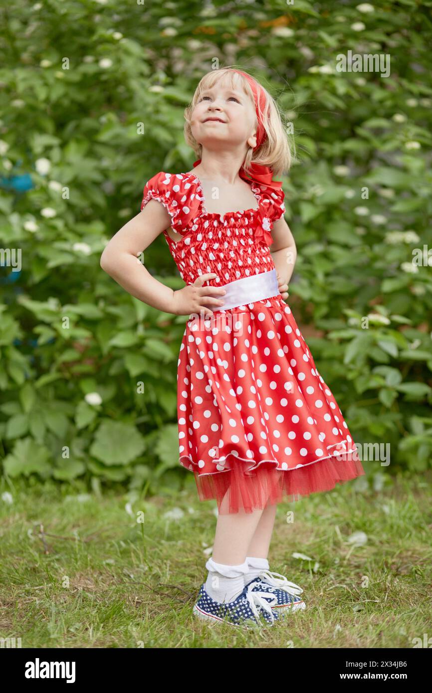 Little girl in red polka-dotted dress stands with  her hands akimbo and looking upward at grassy lawn. Stock Photo