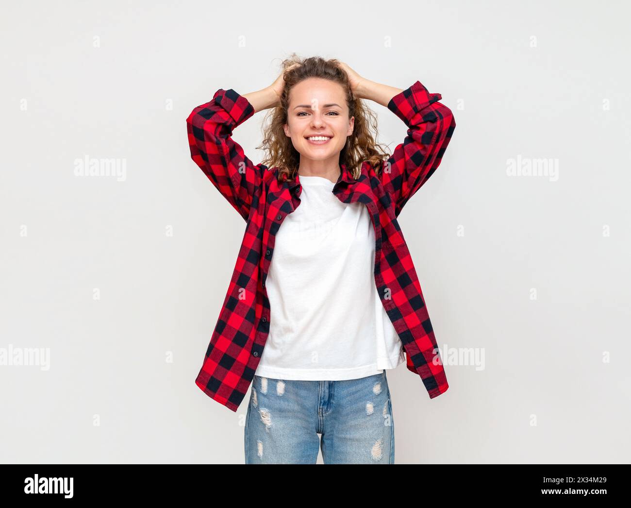 Happy woman holds hands behind her head smiles expressing delight anticipation emotions in front of light plain background in studio. Stock Photo
