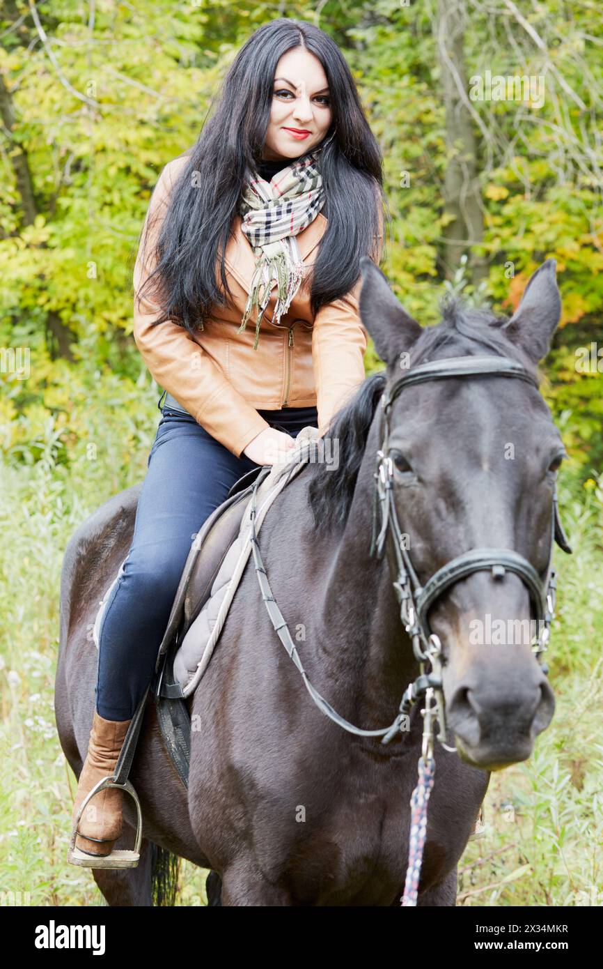 Black-haired woman in brown jacket sits on bay horse in park. Stock Photo