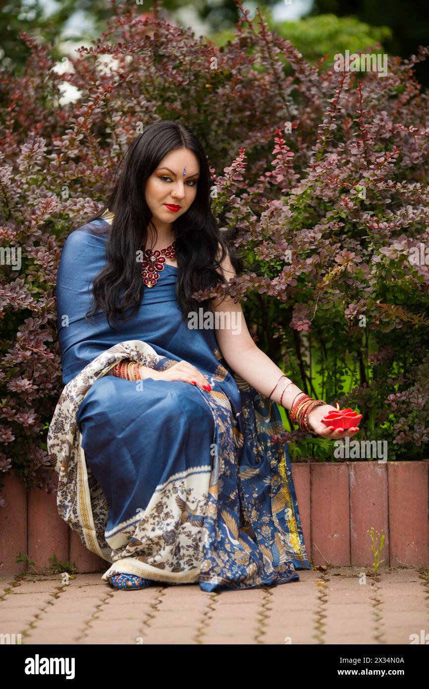 Brunette in blue sari sits near barberry bushes with red burning candle in form of roses Stock Photo