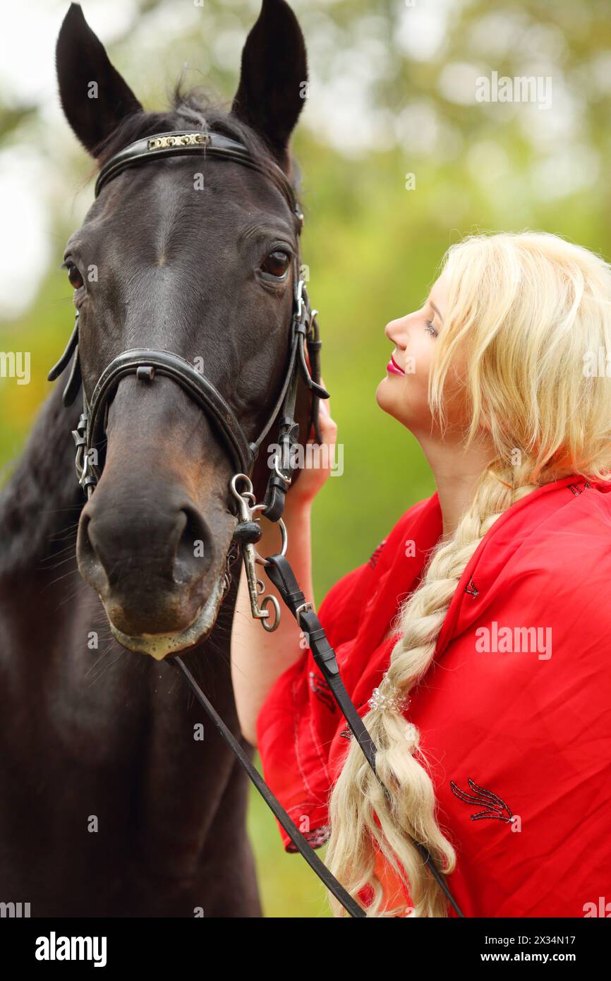 Woman in red dress and long blond hair strokes head of black horse in park Stock Photo