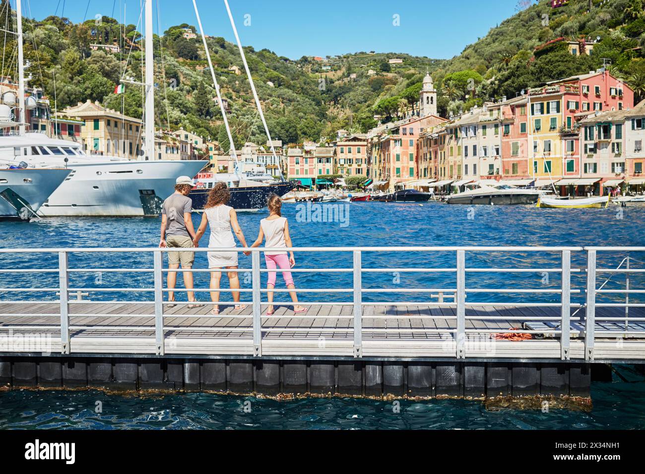 Woman and two children stand holding hands and looking at boats in sea bay with colorful buildings on the shore. Stock Photo