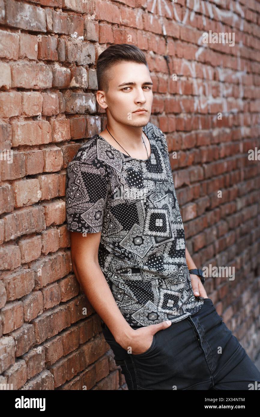 Young Hipster Guy In A Black Shirt Standing Near Red Brick Wall With Graffiti. Stock Photo