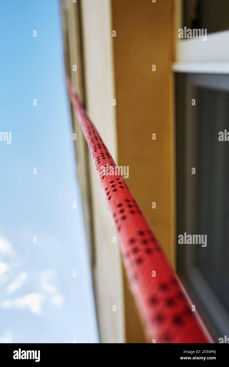 Red industrial climbing rope hanging down from house roof along wall. Stock Photo