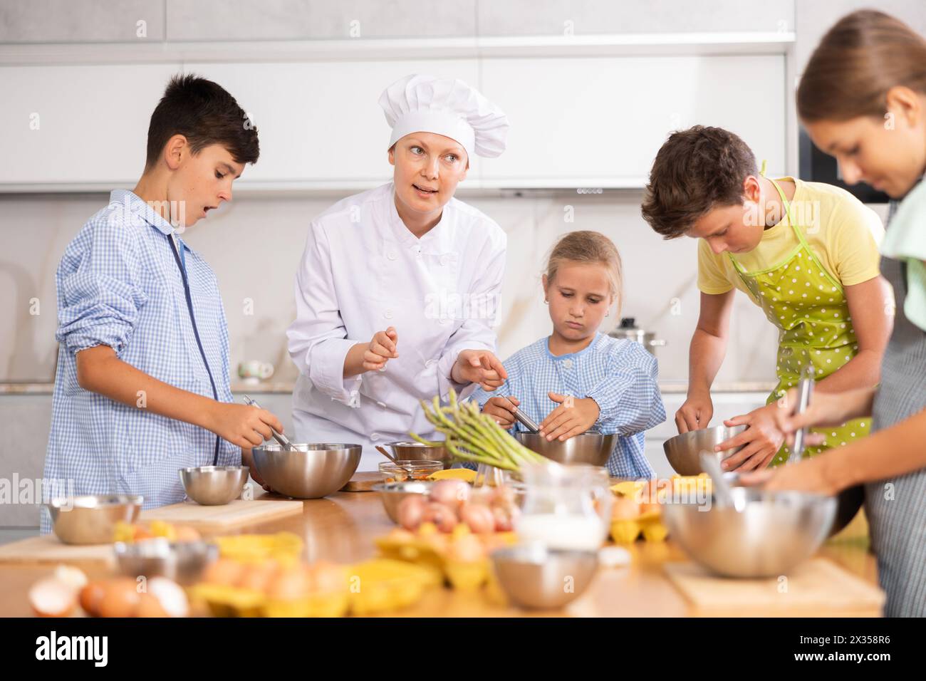Friendly female chef in white uniform giving culinary lesson to tweens ...