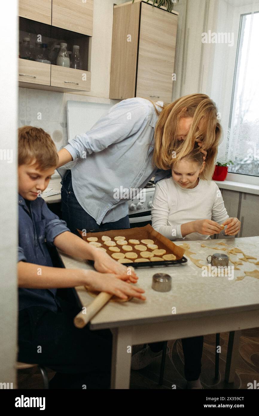 A woman and two children are gathered around a kitchen counter, rolling out dough and cutting cookies into shapes. They are wearing aprons and surroun Stock Photo