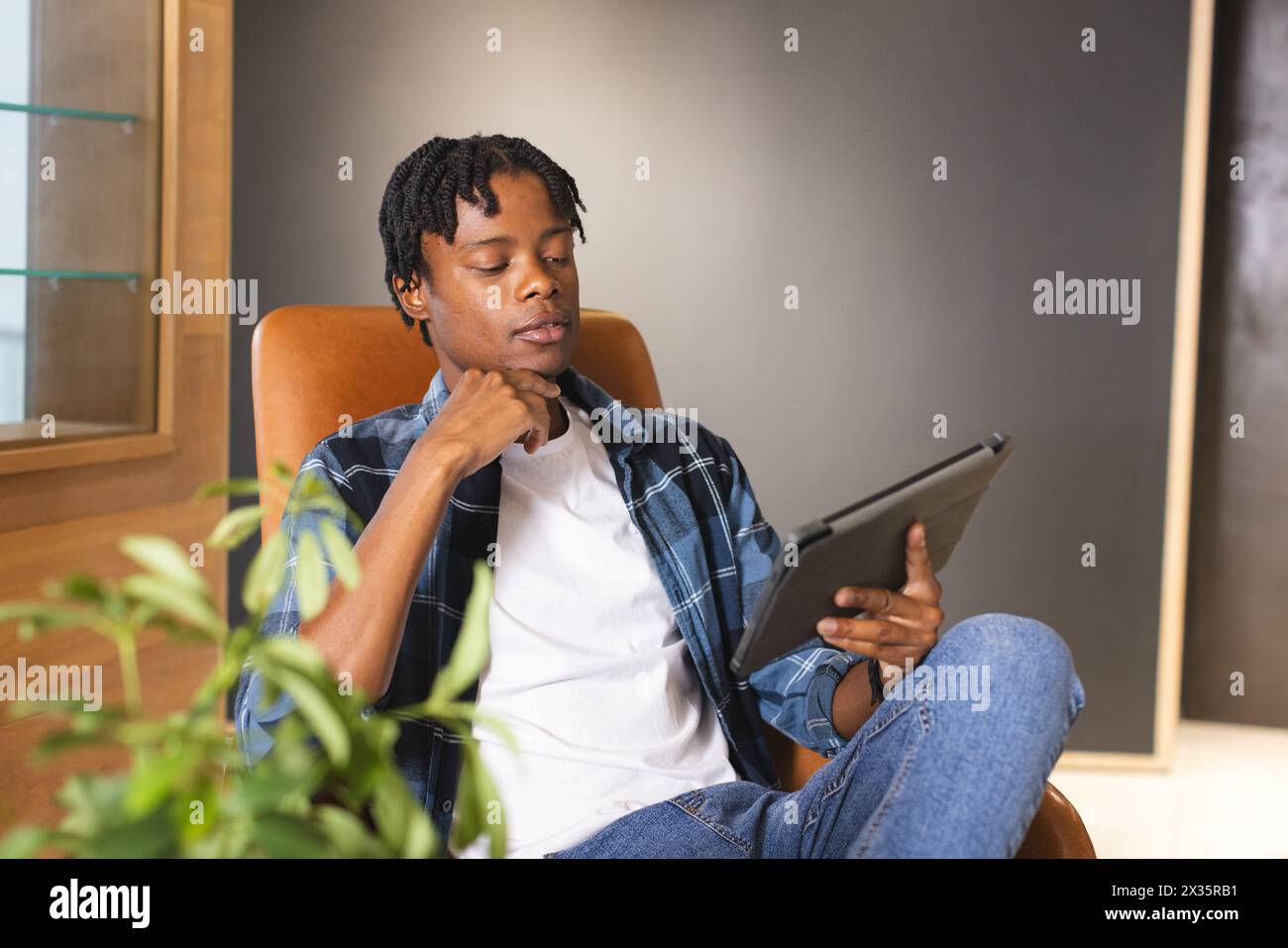 African American man holding tablet, sitting in chair, looking thoughtful in a modern business offic Stock Photo