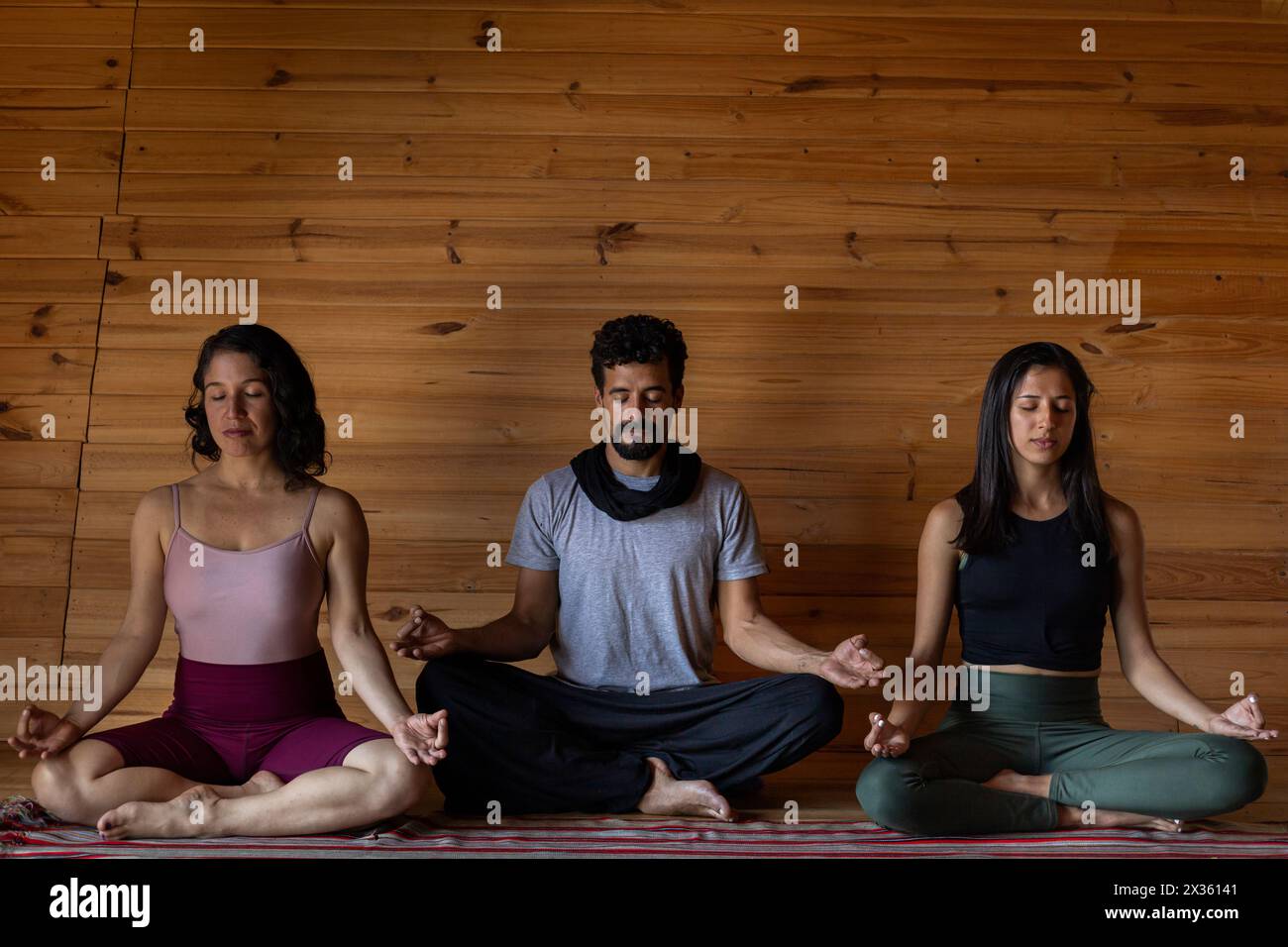 Front view of three young Latin Americans meditating together in yoga class. Background of wooden slats. Concept of wellness and yoga. Stock Photo