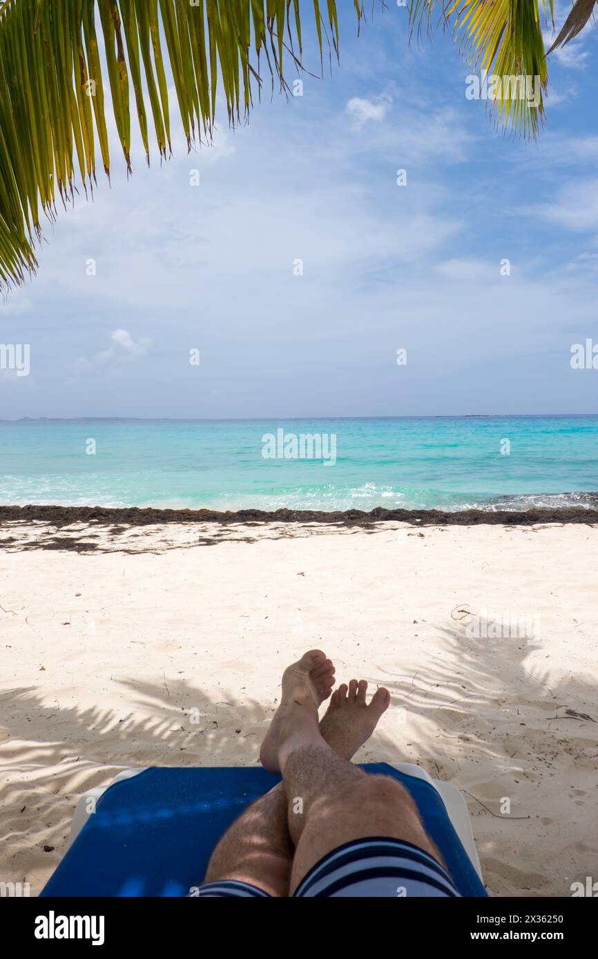 A man is laying on a blue beach chair with his feet up on the sand. The beach is calm and peaceful, with the ocean in the background. The man is enjoy Stock Photo