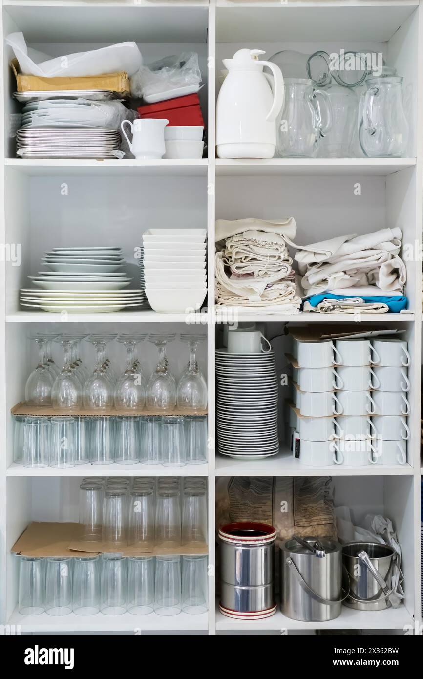 A white cabinet with many dishes and glasses on the shelves. The cabinet is full of various items, including cups, plates, and bowls. Scene is clutter Stock Photo