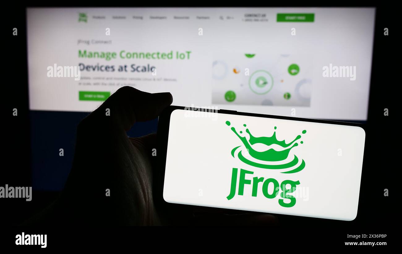 Person holding smartphone with logo of US software development company JFrog Ltd. in front of website. Focus on phone display. Stock Photo