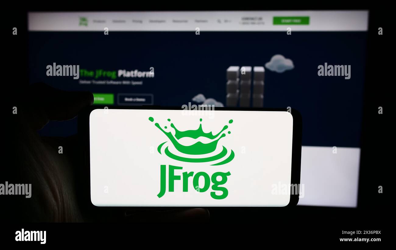Person holding cellphone with logo of US software development company JFrog Ltd. in front of business webpage. Focus on phone display. Stock Photo