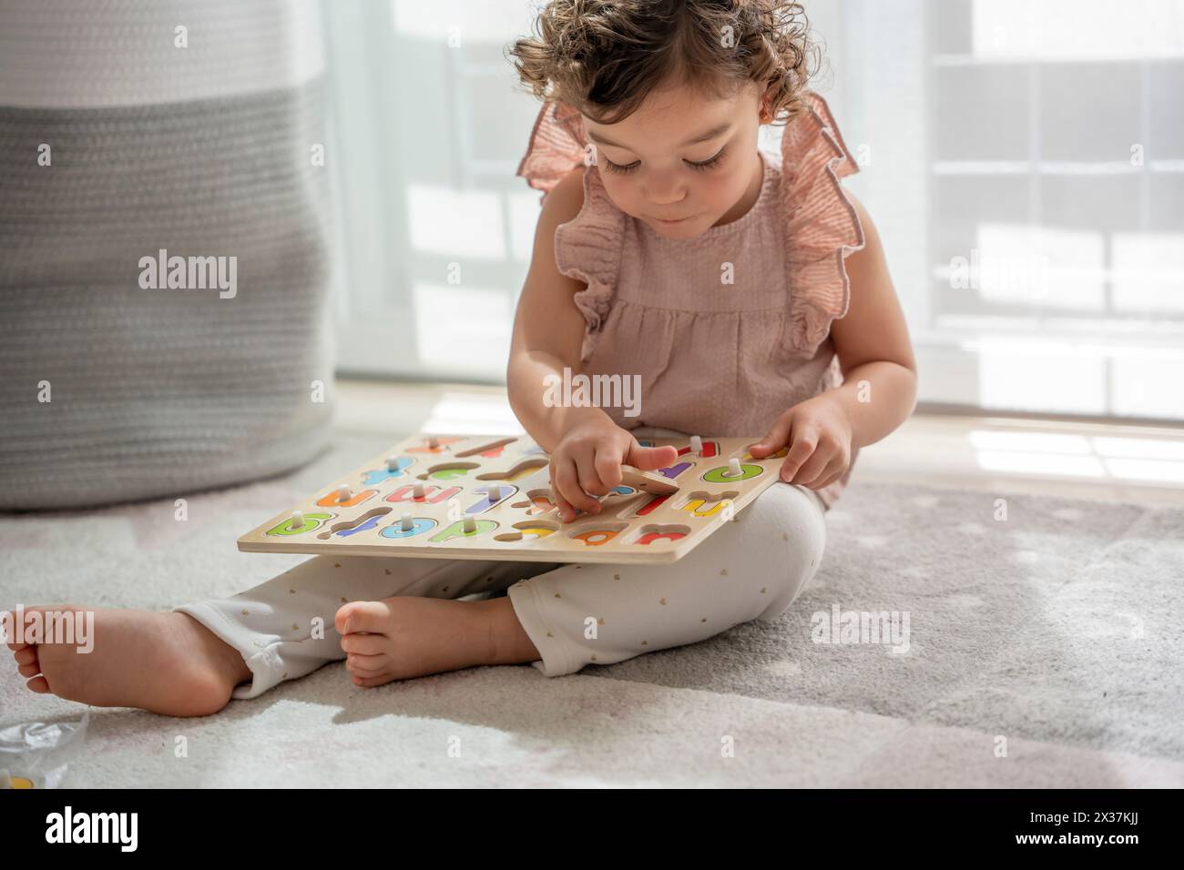 horizontal image of a little girl with curly hair playing sitting on the carpet in her room with a wooden alphabet, Montessori material. concept devel Stock Photo