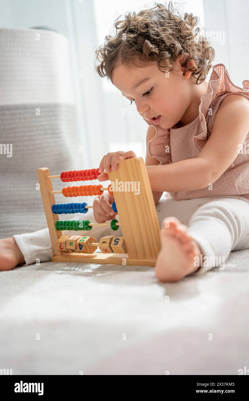 little girl with curly hair playing sitting on the carpet in her room with a wooden toy made of beads, numbers and colors. montessori material concept Stock Photo
